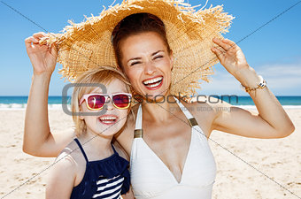Smiling mother and daughter under big straw hat at sandy beach