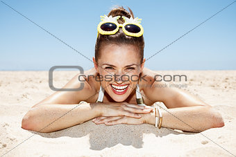 Smiling woman in swimsuit and pineapple glasses laying on beach
