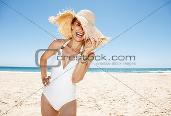 Smiling woman in swimsuit hiding in big straw hat at sandy beach