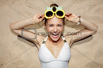 Cheerful woman in swimsuit and pineapple glasses laying on sand