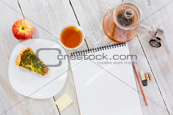 Drawing-pad, cup of tea, chicken pie, apple, glass teapot and watch on wooden table