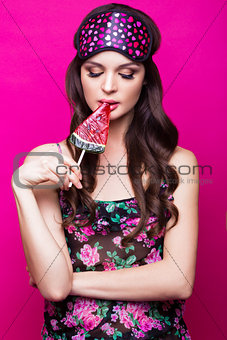 Funny young woman in sleeping mask and pajamas, sweets on pink background. beauty face.