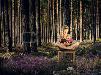 Beautiful blond girl sitting on a chair and reading book in the forest.