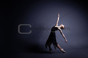 Young ballerina in a black suit is dancing in dark studio