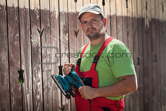 Male worker with vibrating sander in front of old wooden fence