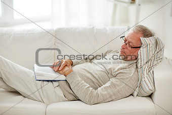 senior man sleeping on sofa with book at home
