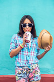 Girl in a city with  cap in shirt drinking  milkshake, fresh juice, enjoying, brunette, tanned, sensual make-up, recreation concept, travel, lifestyle. Summer day. Against the backdrop of  blue wall  red lipstick.