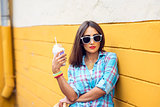 Woman in the city on the background of a yellow wall, holding  milkshake, fresh juice in glasses, wearing  shirt with the bracelets  her arm, makiyah sensual red lips, recreation concept lifestyle. Happy enjoying.