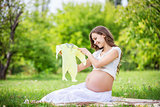 Beautiful pregnant woman holding baby bodysuit and smiling outside in the park