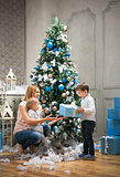 Young mother giving a present to the oldest son, baby boy touching bauble on Christmas tree