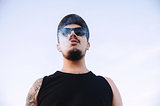 Close-up, portrait of young guy wearing a cap sunglasses with beard and mustache in tattoos, exhaling smoke from cigarette