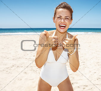 Smiling woman in white swimsuit at sandy beach showing thumbs up