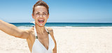 Smiling woman in white swimsuit taking selfie at sandy beach