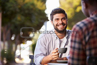 Two adult male friends sit talking over coffee outside cafe