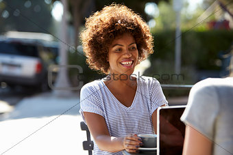 Two female friends with laptop sitting at table outside cafe