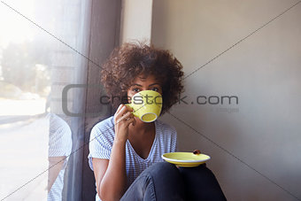 Young black woman sitting by window in cafe drinking coffee