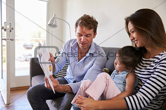 Parents Reading With Baby Daughter On Sofa At Home