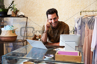 Young man working at clothes shop on phone, using computer