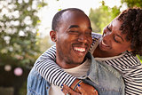 Smiling young black couple piggyback in garden, eyes closed