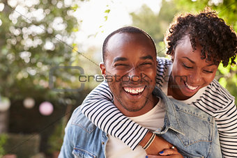 Laughing young black couple piggyback in garden, to camera