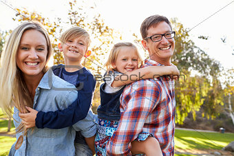 Parents carrying their two young kids piggyback in a park
