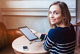 Young female student is enjoying free time,while is sitting with touch pad in coffee shop indoor. Beautiful woman is holding digital tablet and looking at camera during rest in cafe