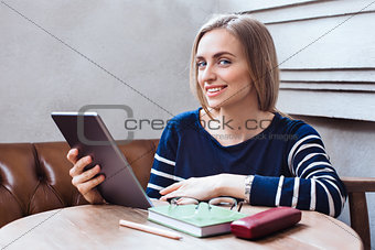 Attractive girl using electronic book on portable touch pad. while is sitting inside the cafe. Smiling and looking at camera. Consept of studing and working online