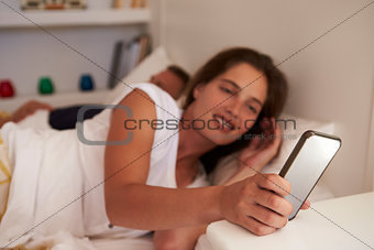Woman in bed uses smartphone, partner sleeps, focus on phone