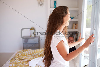 Woman opening bedroom window, her partner in bed, side view