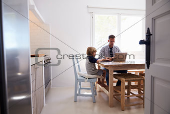 Dad and son using computers at kitchen table, from doorway