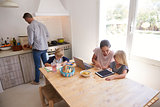 Dad cooking and mum with kids at kitchen table, high angle