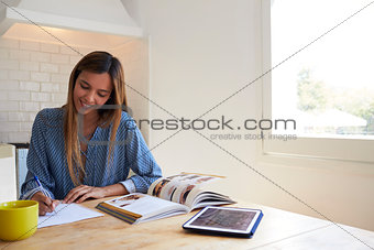 Woman using book and tablet writing in kitchen, close up