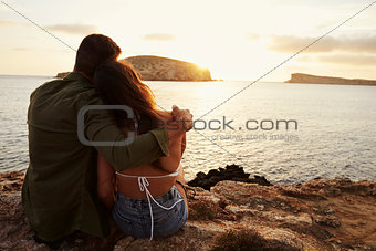 Rear View Of Couple Sitting On Cliff Watching Sunset