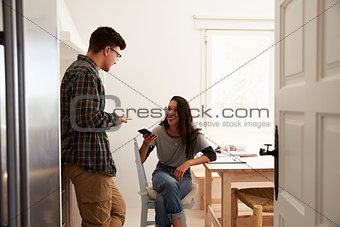Two teenage friends using smartphones, talking in kitchen
