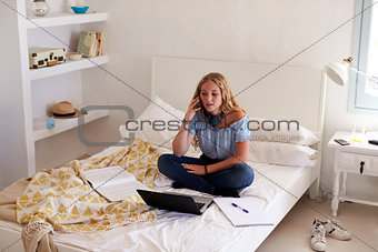 Teenage girl on phone doing homework on her bed with laptop