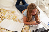 Girl lying on bed using smartphone and laptop, high angle