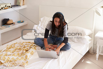 Teenage girl wearing headphones sitting on bed using laptop