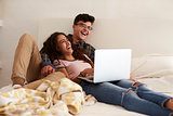 Laughing teenage couple hanging out in bedroom using laptop