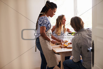 Woman prepares food and friends pour wine, view from doorway