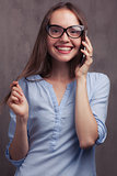 portrait of smiling woman with glasses speaking by cellphone near grey background wall