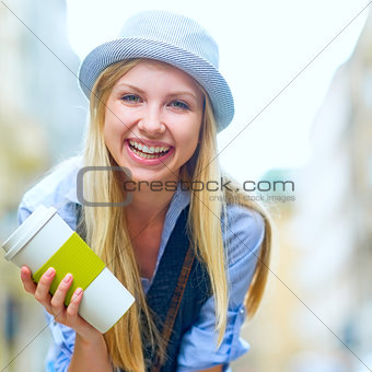 Smiling hipster girl with cup of hot beverage on city street