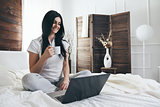 Relaxing on the bed. Beautiful woman enjoying a cup of coffee and using her laptop