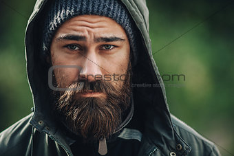 Outdor porttrait of a handsome brutal bearded man with dark beard and mustache dressed in winter clothes, covered with frost. Man wear coat hat and hood. look like hipster.
