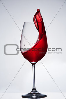 Splashing and moving red wine in the pure wineglass standing against light background with reflection on the stand.