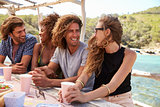 Four friends sitting at a table by the sea look to camera