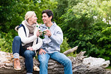 Lovers in vacation sitting at waterside clinking beer bottles