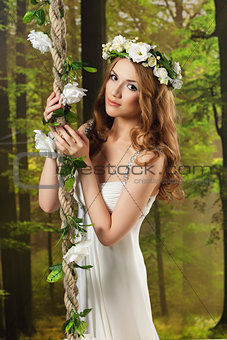 Young girl in a long white dress, with a wreath on a swing in the studio
