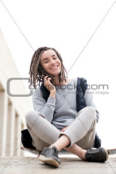 Attractive and smiling teenager girl relaxing with a skateboard and sitting down, talking to somebody on phone