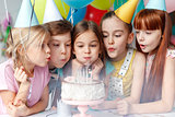 Festive children in party caps, blow candles on delicious cake, make wish, celebrate birthday, have party together, hold colourful balloons. Happy small girl spends festive event with best friends