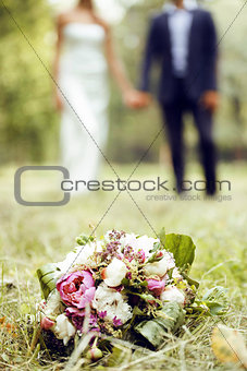 wedding flowers bouquet laying on green grass in park, bride and groom blurred on background holding hands, lifestyle people concept